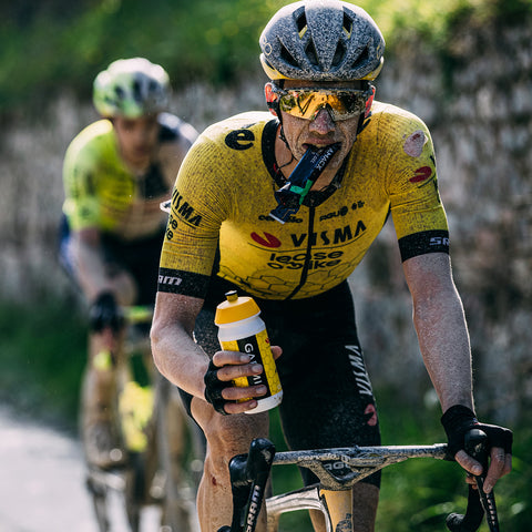 Two cyclists in yellow and black Visma team jerseys ride along a road. The front cyclist, dirt-covered, drinks from a water bottle while holding an Amacx Turbo Gel energy packet with his teeth—a perfect example of maintaining energy supply. Both wear helmets and sunglasses. The background is blurred, indicating movement.