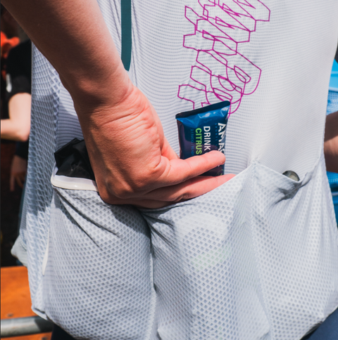 A person places an Amacx Drink Gel | 12-pack packet into the back pocket of a white athletic top adorned with a purple design and multiple mesh pockets, highlighting its fitness-oriented design. The background is slightly blurred, drawing focus to the person's action.
