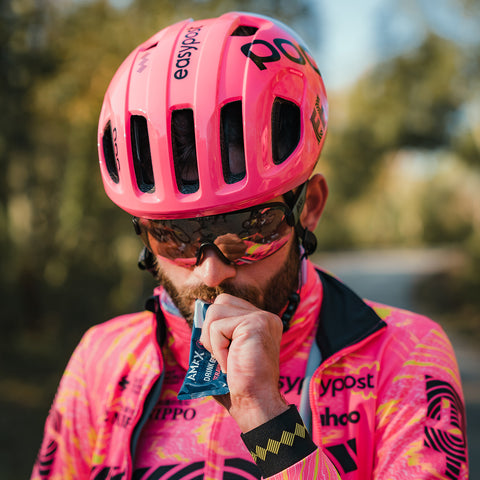 A cyclist, sporting a vibrant pink helmet and matching cycling jersey, stands outside taking a sip from an Amacx Drink Gel | 12-pack. They are wearing sunglasses, with a blurred natural setting of trees and a pathway in the background.