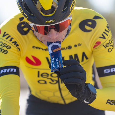 A cyclist in a yellow and black jersey, adorned with sponsor logos from Visma and SRAM, wears sunglasses and a helmet as they consume an Amacx Drink Gel from a packet. The background is blurred to highlight the athlete's precise carbohydrate ratio intake.