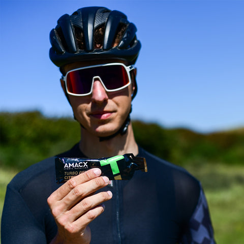 A cyclist wearing a black helmet and white sunglasses holds out an Amacx Turbo Gel from a 12-pack for energy supply. The cyclist, dressed in a black cycling jersey, stands outside on a sunny day with a blurred natural background.