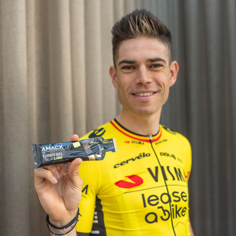 A cyclist in a yellow and black jersey smiles as they hold up an Amacx Turbo Gel from a 12-pack in the Turbo Lime flavor. Ideal for energy supply, this sports gel ensures a steady flow of carbohydrate sources. The background features a neutral-colored curtain.