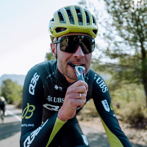 A cyclist wearing a green helmet, black and green cycling suit, and large sunglasses is riding a bike while enjoying an Amacx Drink Gel from a 12-pack. The background features a road lined with trees and another cyclist in the distance. The weather appears to be clear and sunny.