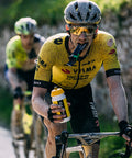 Two cyclists in yellow and black Visma team jerseys ride along a road. The front cyclist, dirt-covered, drinks from a water bottle while holding an Amacx Turbo Gel energy packet with his teeth—a perfect example of maintaining energy supply. Both wear helmets and sunglasses. The background is blurred, indicating movement.