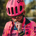 A cyclist wearing a bright pink helmet and matching jersey adorned with various brand logos, including Amacx.com’s Giro d'Italia Package, enjoys an Energy Drink Forest Fruit from a small packet. With sunglasses and a beard, he stands outdoors against a blurred backdrop of lush greenery.