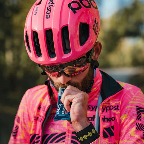 A cyclist wearing a bright pink helmet and matching jersey adorned with various brand logos, including Amacx.com’s Giro d'Italia Package, enjoys an Energy Drink Forest Fruit from a small packet. With sunglasses and a beard, he stands outdoors against a blurred backdrop of lush greenery.