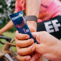 A close-up shot captures a cyclist in motion wearing a pink and white jersey with a black armband, as they eagerly receive an energy chew from the Amacx Endurance Bundle by Amacx.com, handed over by another person. The bike handlebars are partially visible in the frame.