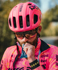 A cyclist, sporting a vibrant pink helmet and matching cycling jersey, stands outside taking a sip from an Amacx Drink Gel | 12-pack. They are wearing sunglasses, with a blurred natural setting of trees and a pathway in the background.
