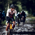 A cyclist wearing a muddy white and red jersey rides through a wet, muddy trail during a race, surrounded by blurred trees. Other cyclists follow in the background, and a photographer crouches by the trail capturing the event.