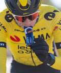 A cyclist in a yellow and black jersey, adorned with sponsor logos from Visma and SRAM, wears sunglasses and a helmet as they consume an Amacx Drink Gel from a packet. The background is blurred to highlight the athlete's precise carbohydrate ratio intake.