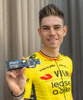A cyclist in a yellow and black jersey smiles as they hold up an Amacx Turbo Gel from a 12-pack in the Turbo Lime flavor. Ideal for energy supply, this sports gel ensures a steady flow of carbohydrate sources. The background features a neutral-colored curtain.