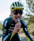 A cyclist wearing a green helmet, black and green cycling suit, and large sunglasses is riding a bike while enjoying an Amacx Drink Gel from a 12-pack. The background features a road lined with trees and another cyclist in the distance. The weather appears to be clear and sunny.